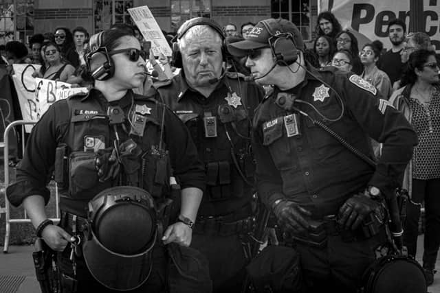 Three police officers wearing headsets stand before a crowd holding protest signs.