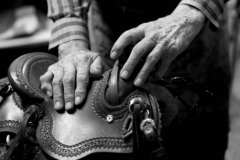 Weathered hands rest on the decorated leather seat and horn of a saddle.