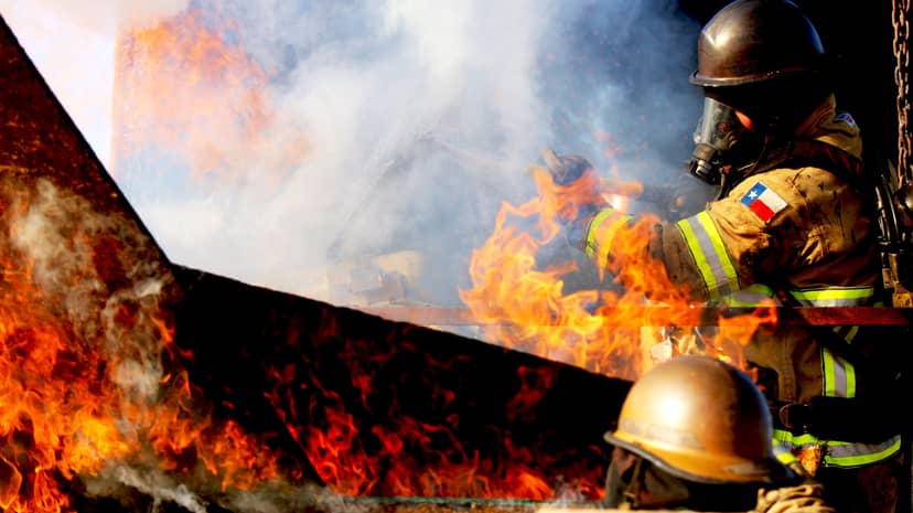 Firefighter in protective gear works amid bright flames and thick smoke.