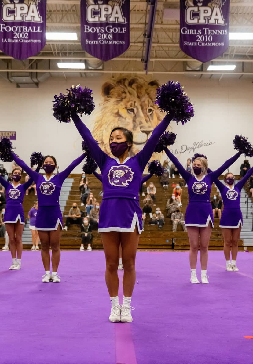 Masked cheerleaders in purple uniforms hold pom-poms overhead on a gymnasium floor.