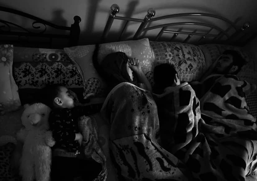 Four people sleep together under patterned blankets in a dark bedroom.