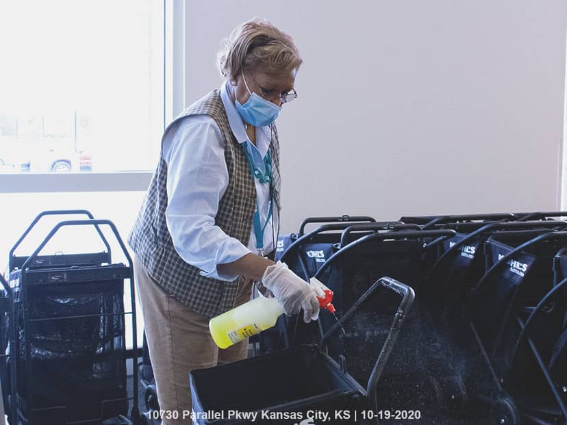 Masked worker wearing gloves sprays disinfectant onto rows of nested shopping carts.