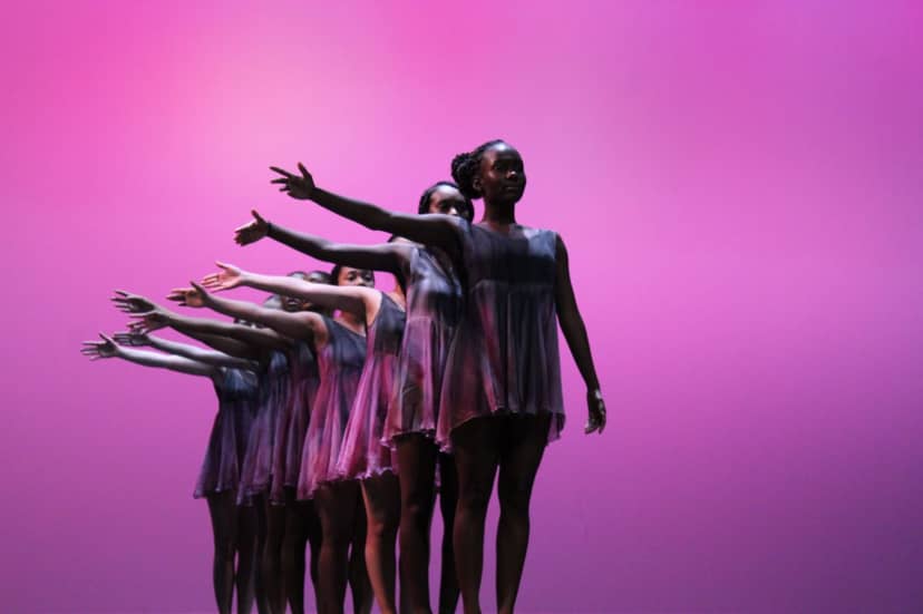 Dancers in matching dresses extend their arms in formation against a pink-lit stage.