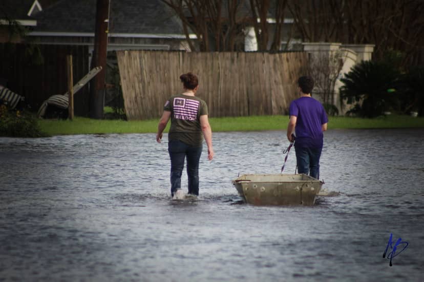 Two people wade through floodwater while pulling a small metal boat toward a fenced yard.