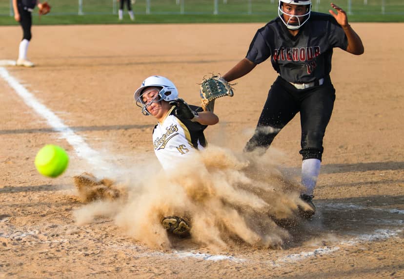Softball player slides into home plate as catcher reaches toward her through billowing dust.