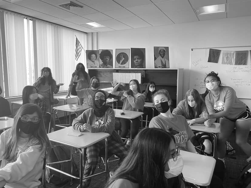 Masked students sit at classroom desks, several raising their arms toward the camera.