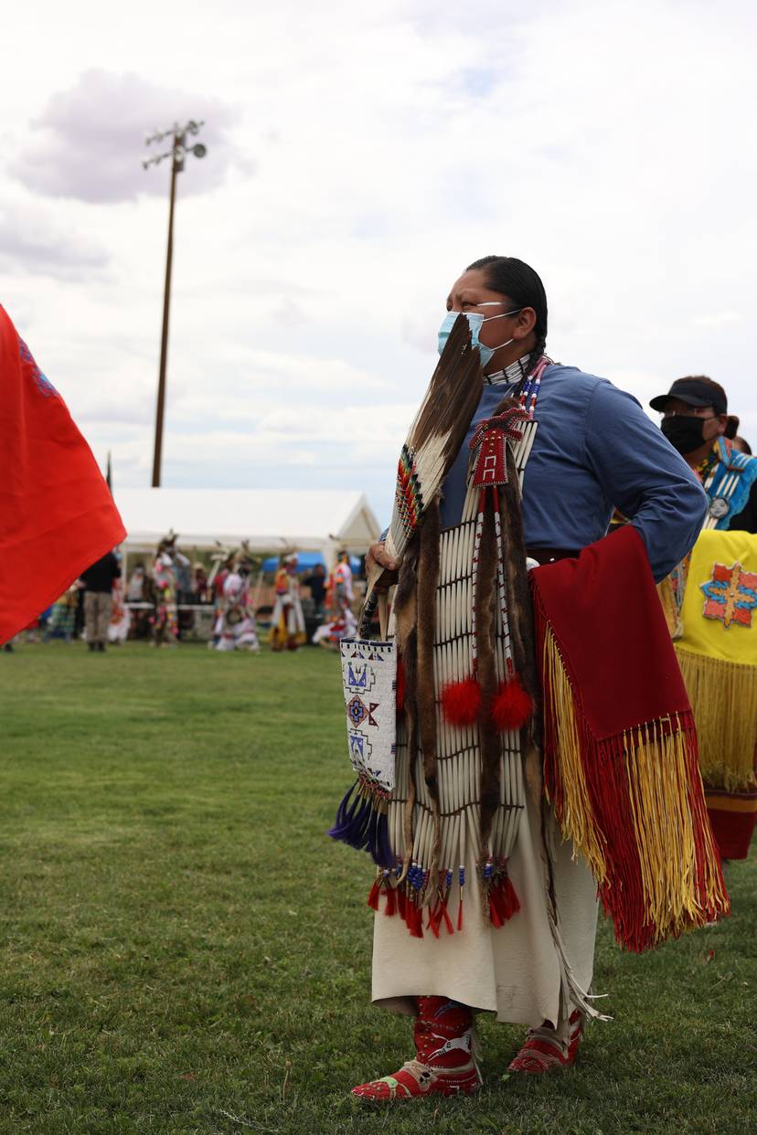 Masked participant wearing feathered regalia stands on a grassy field during an outdoor gathering.