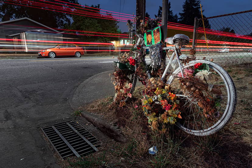 Flower-covered white memorial bicycle rests beside a road streaked by passing car lights.