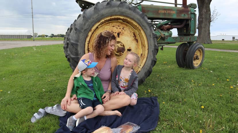 Adult and two children sit on a picnic blanket beside an antique tractor.