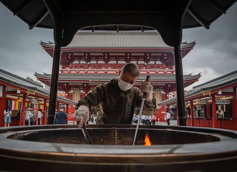 Masked worker tends a smoking incense burner before an ornate red temple gate.