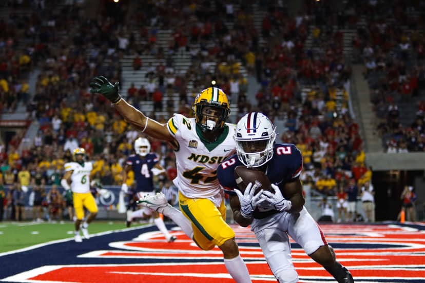 Football receiver cradles the ball in the end zone as a defender reaches.