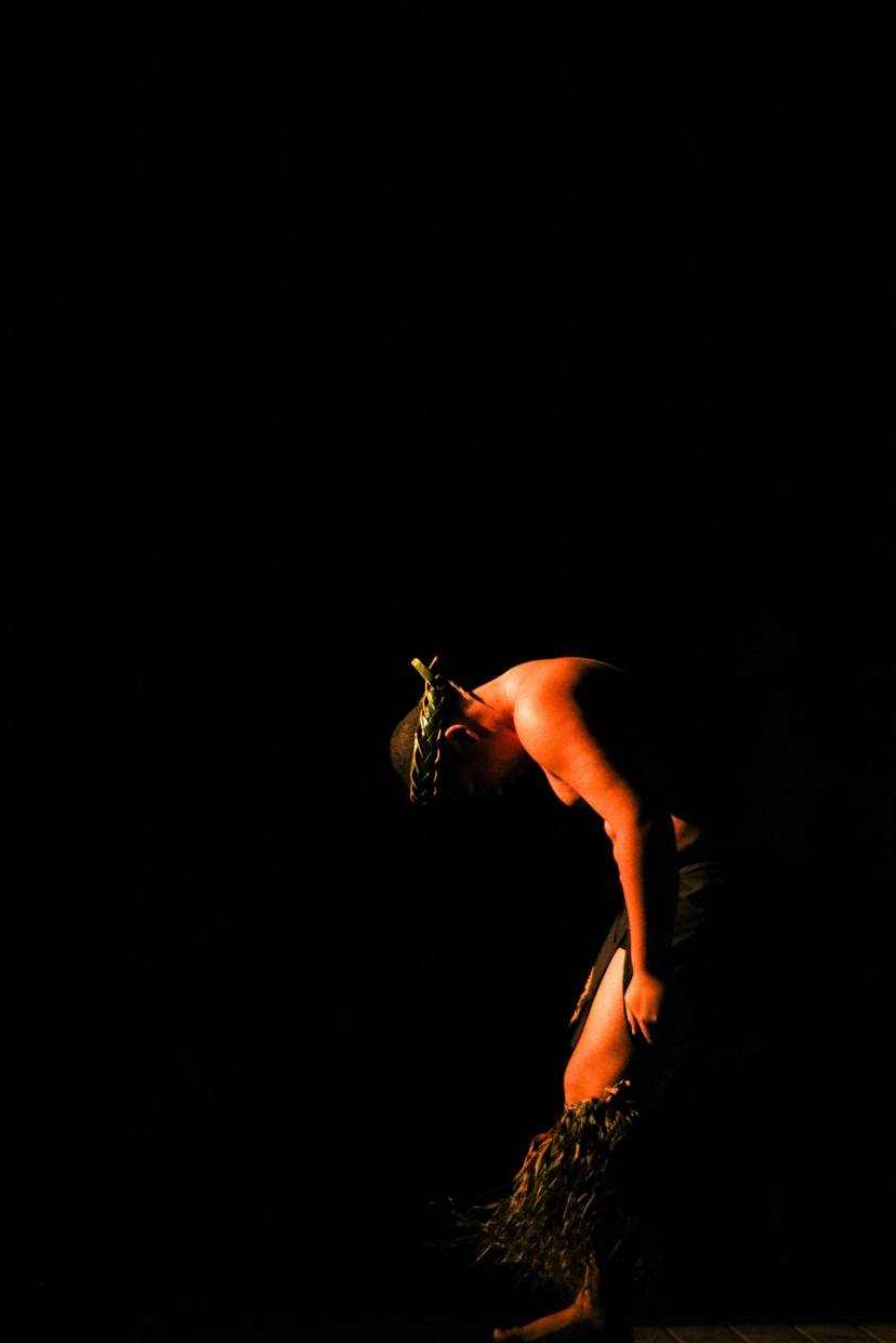 Spotlit performer bends forward against a black background in a textured costume.