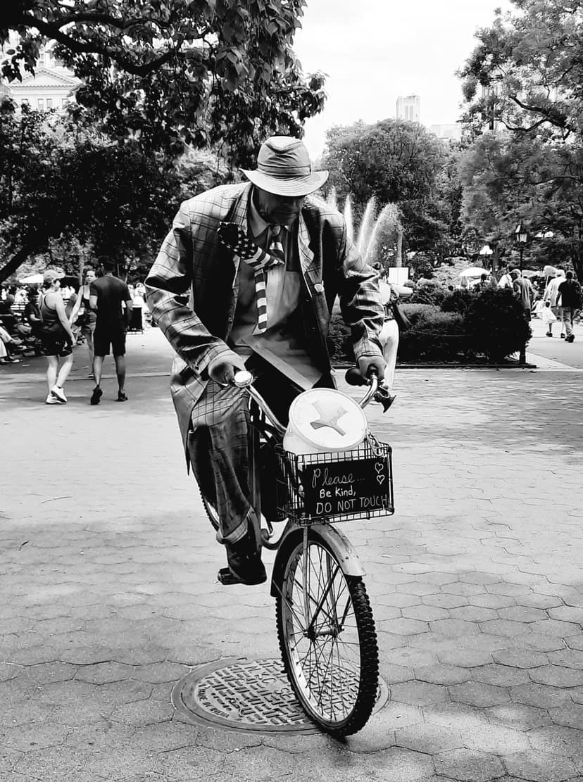 Suited cyclist rides through a park with an instruction sign on the basket.