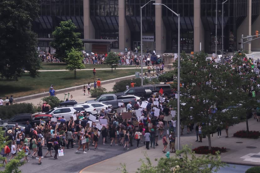 Large protest crowd carrying signs fills a city street outside a government building.