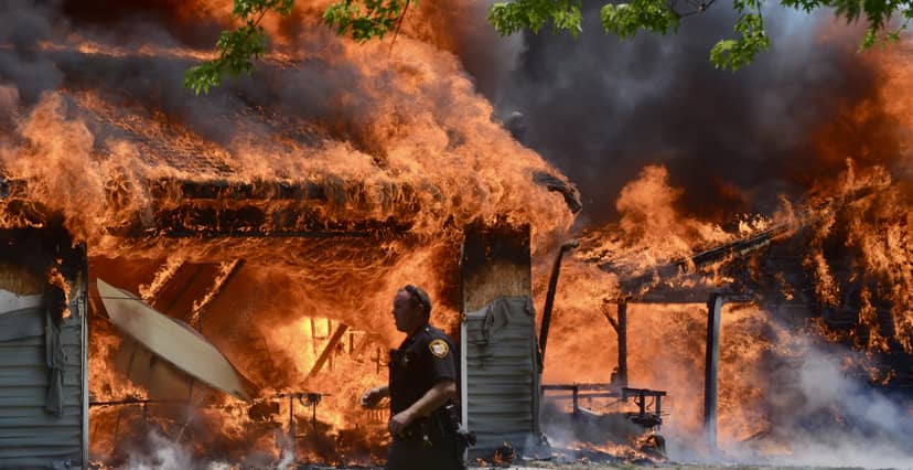 Police officer walks before a house engulfed in flames and smoke.