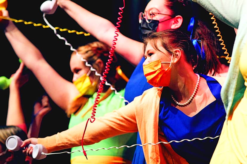 Masked performers in colorful costumes hold telephone receivers amid coiled cords.