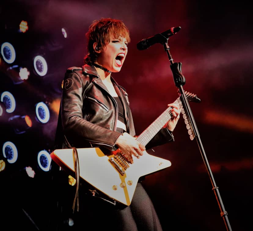 Musician sings into a microphone while playing guitar under red stage lights.