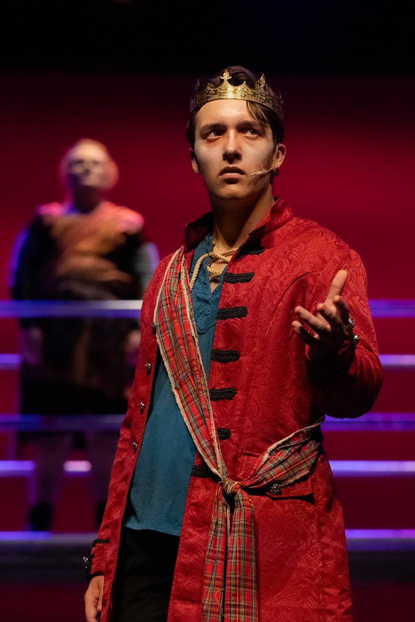 Crowned stage actor in a red coat gestures beneath red lighting.