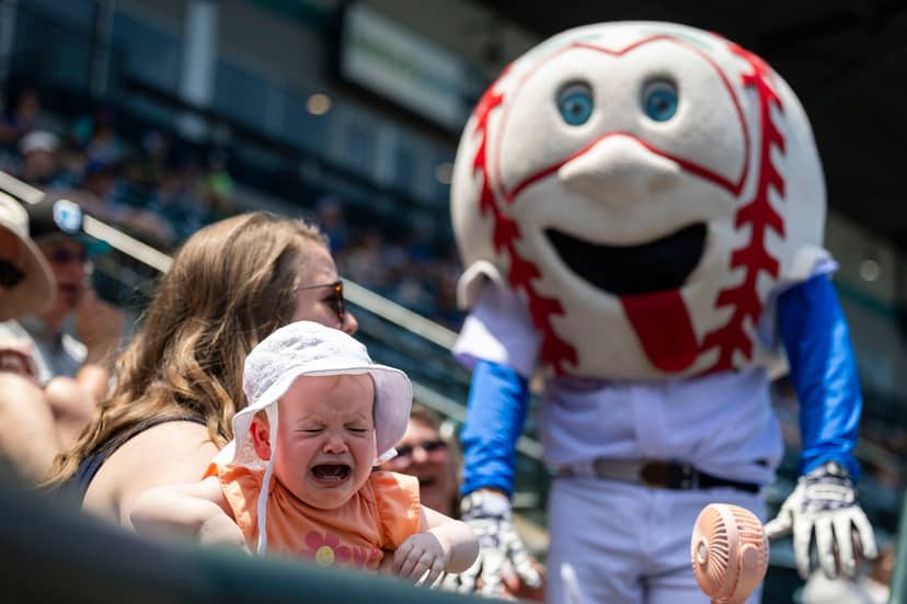 Crying baby sits near a smiling baseball mascot and stadium spectators.