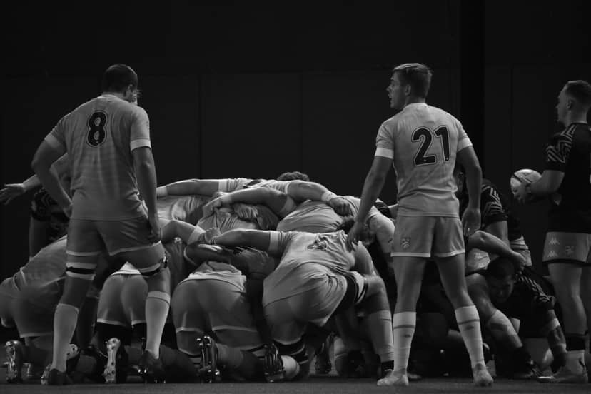 Rugby players form a scrum as teammates and an official stand nearby.