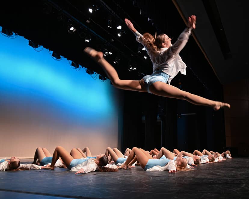 Dancer leaps above a row of performers lying across a theater stage.