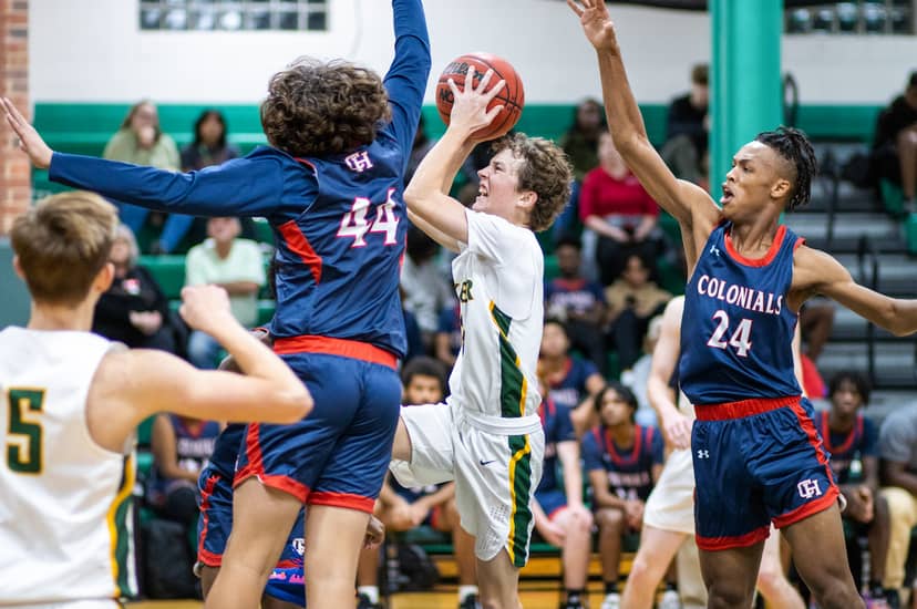 Basketball player drives toward the basket between two defenders with raised arms.