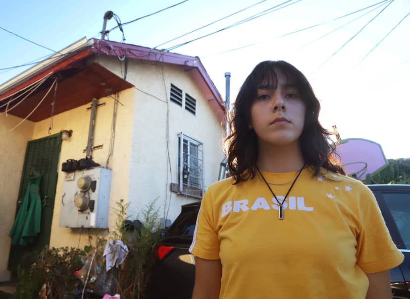 Person in a yellow Brasil shirt stands before a house, cars, and basketball hoop.