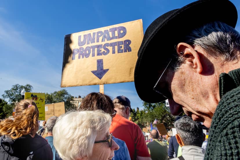 Crowd gathers beneath an Unpaid Protester sign as two people converse in the foreground.