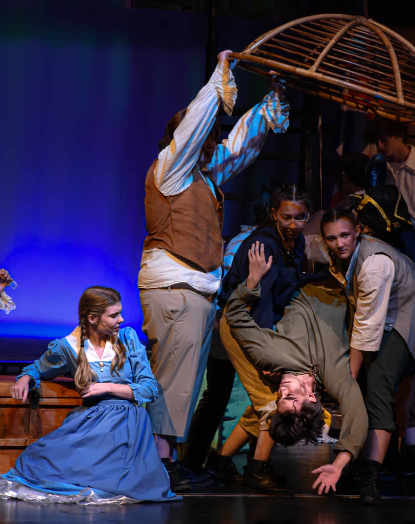 Costumed stage performers cluster beneath a wooden cage as one hangs upside down.